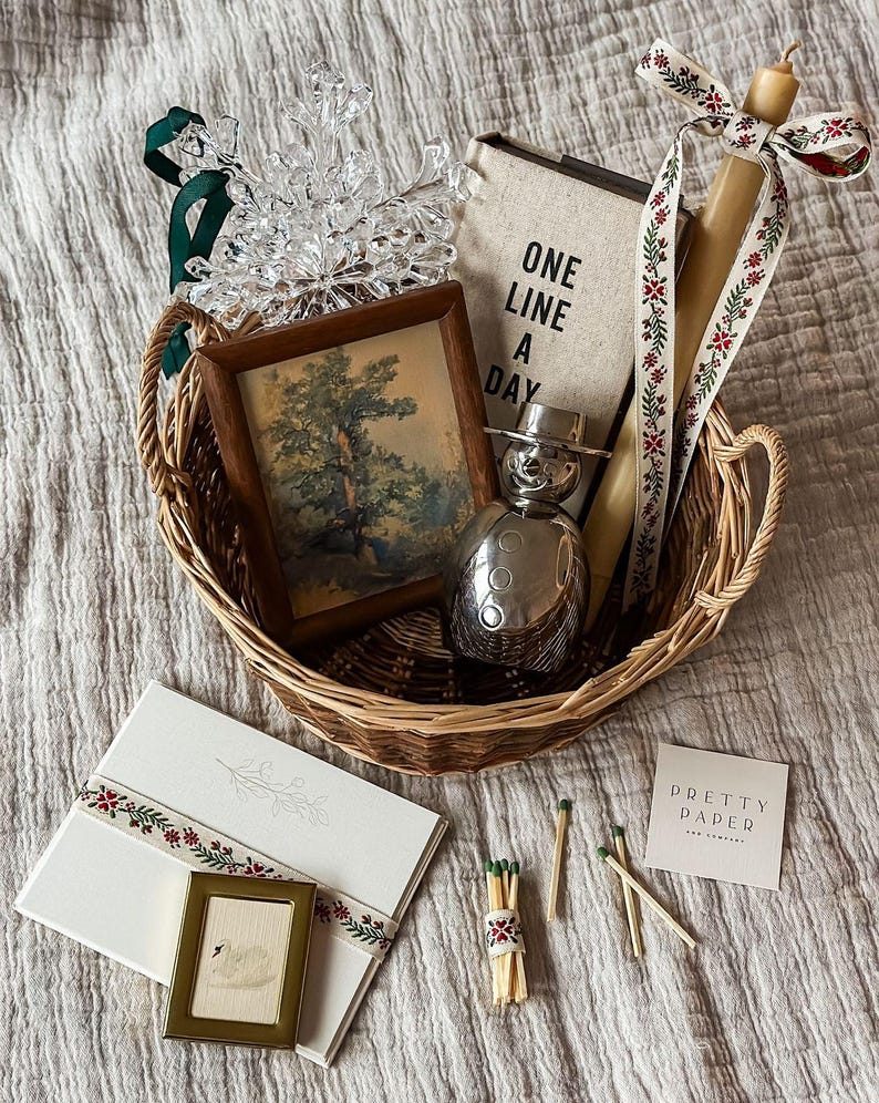 May include: A woven basket filled with decorative items. Includes a framed landscape painting, a journal with "ONE LINE A DAY" text, a snowman-shaped perfume bottle, a candle with a floral ribbon, a snowflake ornament, and stationery with a small framed picture.