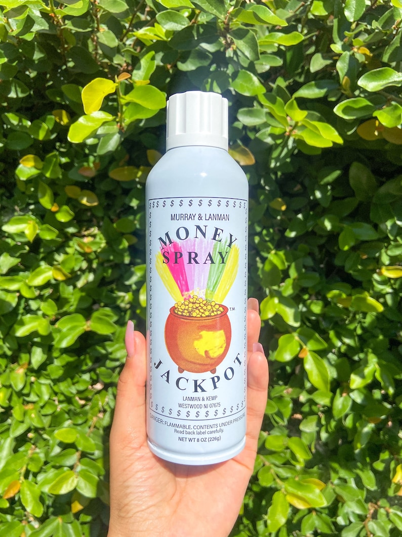 May include: A light blue spray bottle with the text "MONEY SPRAY JACKPOT" and a pot of gold illustration. The bottle is held in front of a green leafy background. The bottle is 8 oz.
