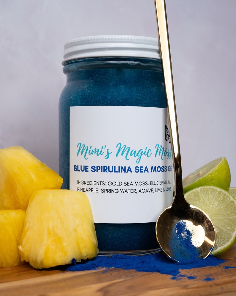 K&ouml;nnte beinhalten: Ein Glasgef&auml;&szlig; gef&uuml;llt mit einem blauen Spirulina-Seetang-Gel. Das Gef&auml;&szlig; hat einen wei&szlig;en Deckel und ein Etikett mit der Aufschrift "Mimi's Magic Moss" und "Blue Spirulina Sea Moss Gel". Die Zutaten sind auf dem Etikett aufgelistet: "Gold Sea Moss, Blue Spirulina, Pineapple, Spring Water, Agave, Lime & Love". Neben dem Gef&auml;&szlig; befinden sich Ananasst&uuml;cke und ein Limettenst&uuml;ck.