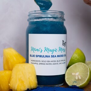 K&ouml;nnte beinhalten: Ein Glasgef&auml;&szlig; gef&uuml;llt mit blauem Spirulina-Seetang-Gel, wobei ein goldener L&ouml;ffel etwas von dem Gel entnimmt. Das Gef&auml;&szlig; hat ein wei&szlig;es Etikett mit dem Text "Mimi's Magic Moss" und "BLUE SPIRULINA SEA MOSS GEL". Die Zutaten sind unten aufgelistet: "GOLD SEA MOSS, BLUE SPIRULINA, PINEAPPLE, SPRING WATER, AGAVE, LIME & LOVE". Auf einer Holzoberfl&auml;che liegen Ananasst&uuml;cke und Limettenscheiben, wobei blaues Pulver um das Gef&auml;&szlig; herum verstreut ist.