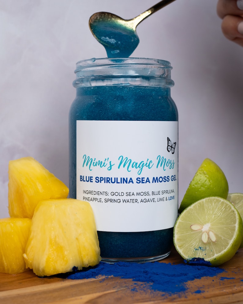 K&ouml;nnte beinhalten: Ein Glasgef&auml;&szlig; gef&uuml;llt mit blauem Spirulina-Seetang-Gel, wobei ein goldener L&ouml;ffel etwas von dem Gel entnimmt. Das Gef&auml;&szlig; hat ein wei&szlig;es Etikett mit dem Text "Mimi's Magic Moss" und "BLUE SPIRULINA SEA MOSS GEL". Die Zutaten sind unten aufgelistet: "GOLD SEA MOSS, BLUE SPIRULINA, PINEAPPLE, SPRING WATER, AGAVE, LIME & LOVE". Auf einer Holzoberfl&auml;che liegen Ananasst&uuml;cke und Limettenscheiben, wobei blaues Pulver um das Gef&auml;&szlig; herum verstreut ist.