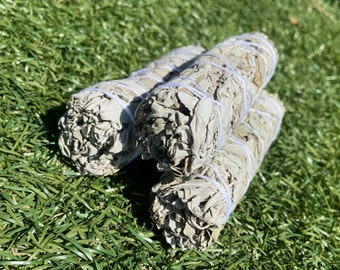 May include: Three bundles of white sage smudge sticks tied with white string. The smudge sticks are on a green artificial grass surface.