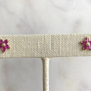 May include: A pair of gold flower stud earrings with pink gemstones. The earrings are displayed on a white fabric stand.