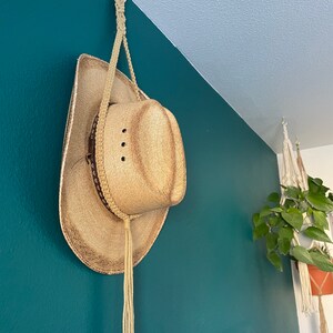May include: A straw cowboy hat with a decorative band and a long tassel hangs from a macrame hanger attached to a white ring. The hat is suspended against a teal wall, with a hanging plant visible in the background.