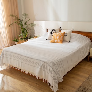 May include: A white bed with a white and gray striped bedspread and a white headboard. The bed has a few decorative pillows with black and white geometric patterns and a yellow pillow with a black design. The bed is in a bedroom with wooden floors and a wooden nightstand.