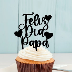 May include: A cupcake with white frosting and a black topper that says "Feliz Dia Papa" with hearts.