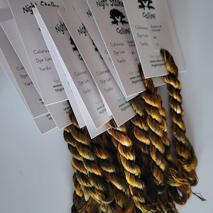 May include: Several skeins of hand-dyed yarn in shades of brown, gold, and green are displayed. Each skein is attached to a white card with the text "Night Stalkers Calling" and other details. The yarn appears to be for crafting.