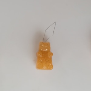 May include: Three orange gummy bear earrings with glitter accents are displayed against a white background. Each bear has a wire hook for pierced ears. The earrings are a fun and playful accessory.
