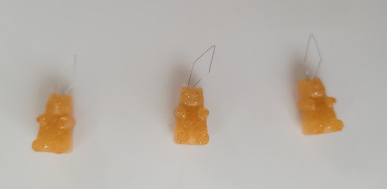 May include: Three orange gummy bear earrings with glitter accents are displayed against a white background. Each bear has a wire hook for pierced ears. The earrings are a fun and playful accessory.