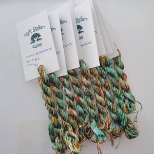 May include: Bundles of colorful embroidery floss with white tags. The tags read "Night Stalkers Calling" with "Bonnie Lass" colorway and "Dye Lot #1". The floss is a mix of green, orange, and blue.