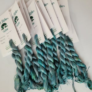 May include: Six skeins of variegated teal and blue embroidery floss attached to white product cards. Each card features the text Night Stalkers Calling, Colorway 3, Dye Lot 10, and web address details. The thread displays a twisted texture.