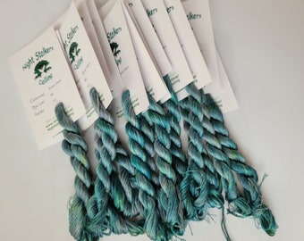May include: Six skeins of variegated teal and blue embroidery floss attached to white product cards. Each card features the text Night Stalkers Calling, Colorway 3, Dye Lot 10, and web address details. The thread displays a twisted texture.