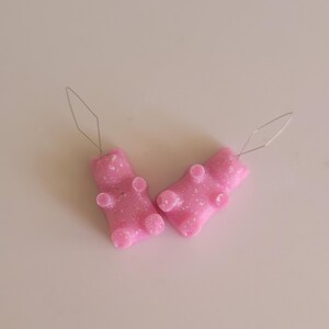 May include: Two pink gummy bear-shaped earrings with silver-colored ear wires. The earrings have a glittery texture and are lying on a white surface. These novelty earrings are a fun accessory.