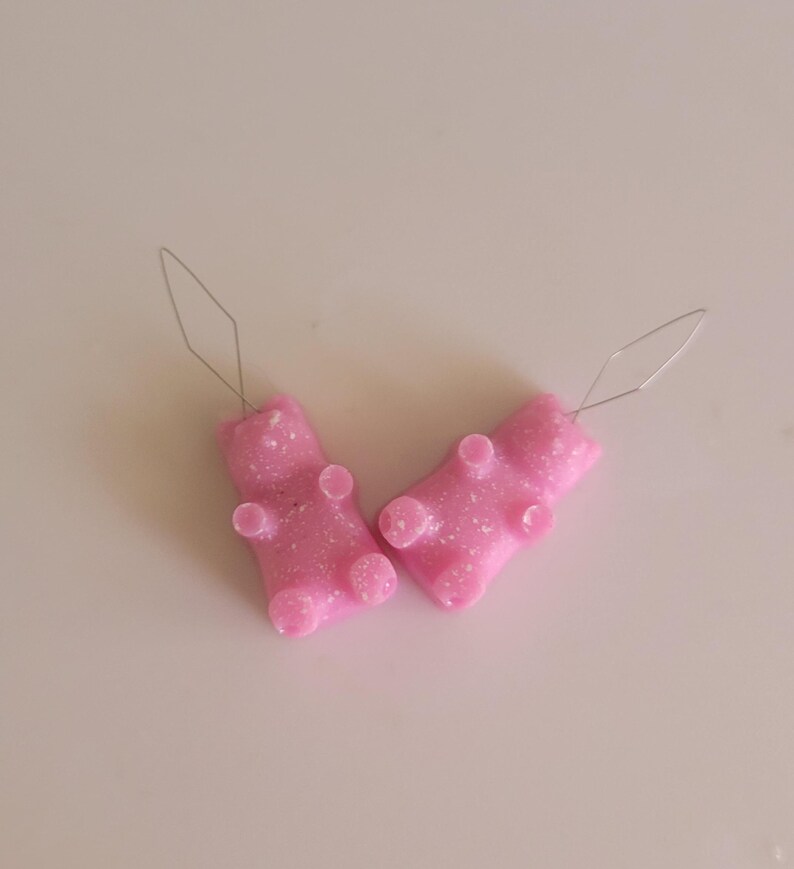 May include: Two pink gummy bear-shaped earrings with silver-colored ear wires. The earrings have a glittery texture and are lying on a white surface. These novelty earrings are a fun accessory.