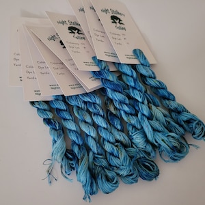 May include: A collection of seven skeins of variegated blue embroidery floss, each attached to a white card labeled Night Stalkers Calling with a frog graphic. The thread features shades of light and dark blue with subtle hints of yellow.