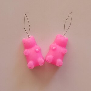 May include: Two bright pink gummy bear-shaped charms with attached silver-colored wire loops. The charms have a smooth, glossy finish and are positioned on a light pink surface. The gummy bears have small details representing arms, legs, and a head.