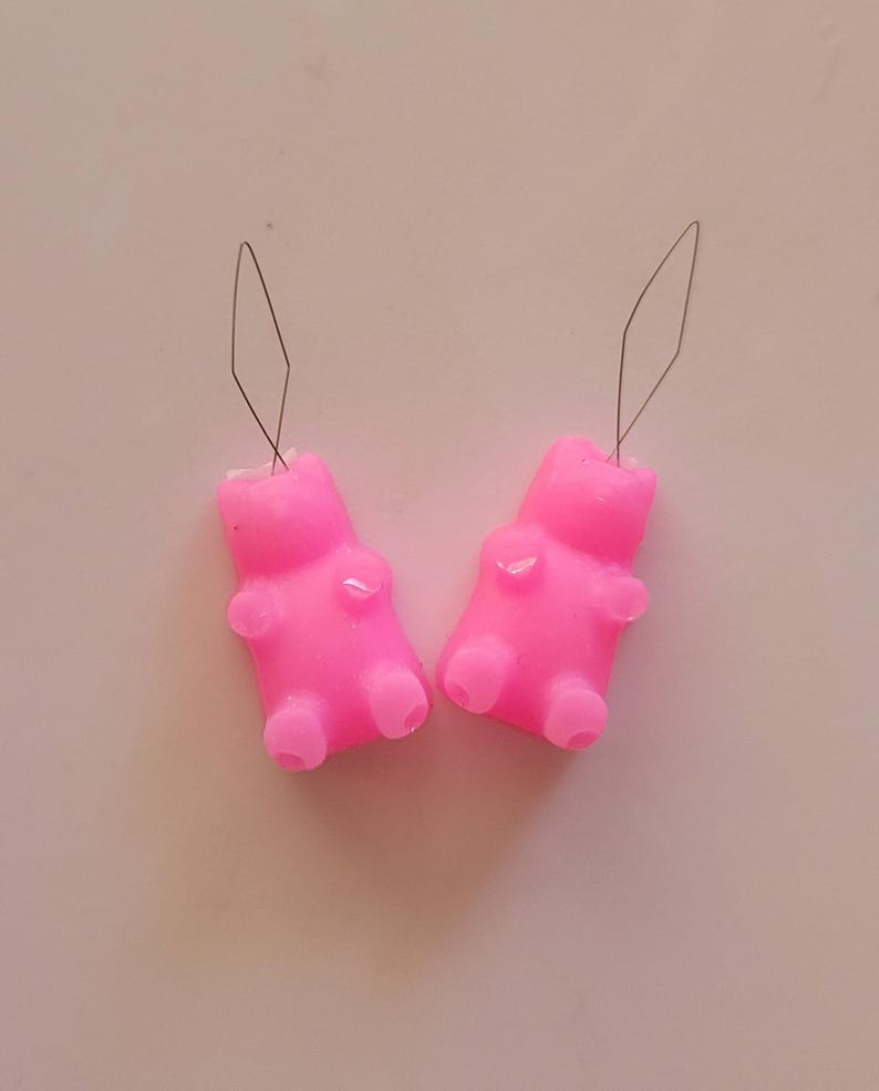 May include: Two bright pink gummy bear-shaped charms with attached silver-colored wire loops. The charms have a smooth, glossy finish and are positioned on a light pink surface. The gummy bears have small details representing arms, legs, and a head.