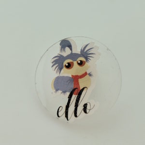 May include: A clear round sticker with a cartoon image of a blue and yellow creature with a red scarf. The creature is wearing a blue scarf and has large eyes. The text "ello" is written in black cursive below the creature.