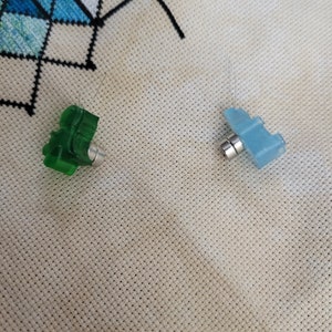 May include: Two small, plastic needle minders. One is green and shaped like a tree, the other is blue and shaped like an airplane. Both have a silver magnet attached to the bottom. The needle minders are on a cream-colored, perforated fabric.