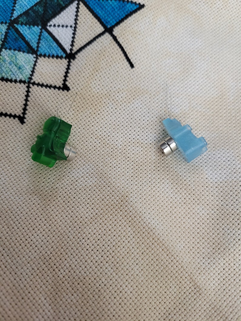 May include: Two small, plastic needle minders. One is green and shaped like a tree, the other is blue and shaped like an airplane. Both have a silver magnet attached to the bottom. The needle minders are on a cream-colored, perforated fabric.