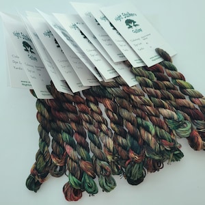 May include: A collection of eight twisted skeins of variegated embroidery thread in shades of dark green, brown, and deep red. Each skein is attached to a white paper tag printed with the text Night Stalkers Calling and product details.