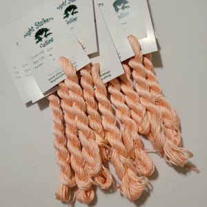 May include: A close-up of eight skeins of peach-colored embroidery floss. The floss is twisted and bundled together. The floss is labeled "Night Stalker's Calling".