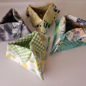 May include: Four fabric origami boxes in various patterns. One is gray with white leaf designs, another is yellow with green leaves, a third is green and white gingham, and the last is teal with floral and leaf patterns.