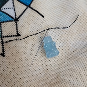 May include: A close-up of a cross-stitch project in progress. A light blue gummy bear-shaped charm is attached to a needle, with black thread. The needle is inserted into the fabric, which has a geometric design in blue and white.