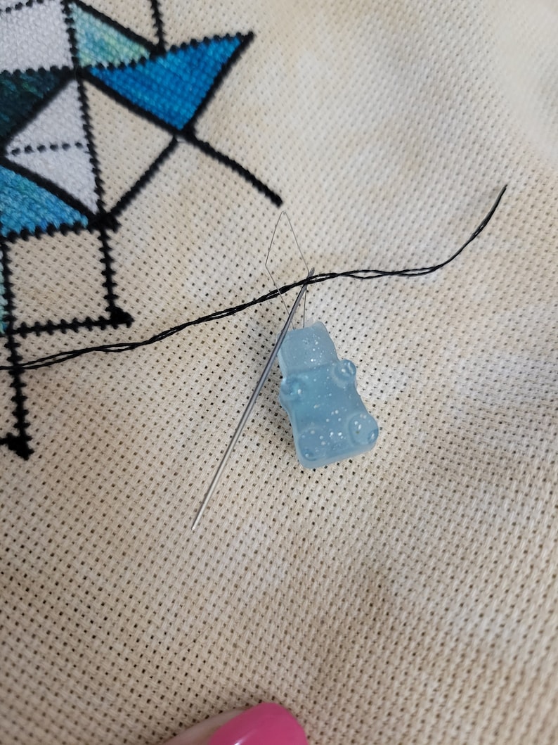 May include: A close-up of a cross-stitch project in progress. A light blue gummy bear-shaped charm is attached to a needle, with black thread. The needle is inserted into the fabric, which has a geometric design in blue and white.