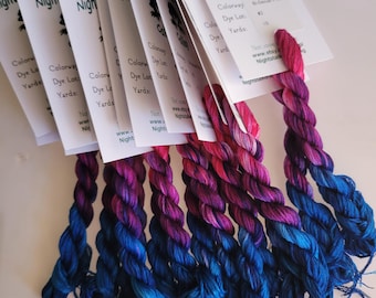 May include: A collection of hand-dyed yarn skeins in shades of blue, purple, and pink. Each skein is attached to a white card with the text "Night Stalkers Calling" and other details. The yarn is arranged in a fan shape.