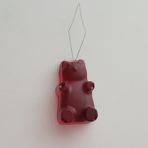 May include: A red gummy bear candy with a wire needle inserted into its head. The gummy bear has a classic bear shape with arms, legs, and a head. The wire needle is bent into a diamond shape.