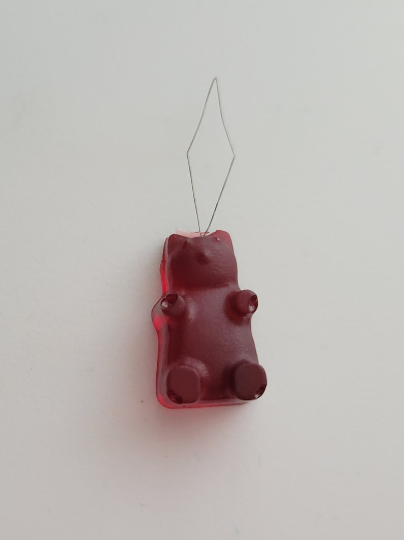 May include: A red gummy bear candy with a wire needle inserted into its head. The gummy bear has a classic bear shape with arms, legs, and a head. The wire needle is bent into a diamond shape.