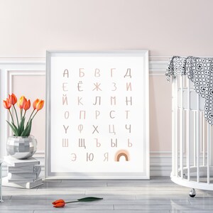 May include: A white framed print with the Cyrillic alphabet in pink and brown letters. The letters are arranged in a grid pattern. A rainbow with pink, orange, and yellow stripes is at the bottom of the print.
