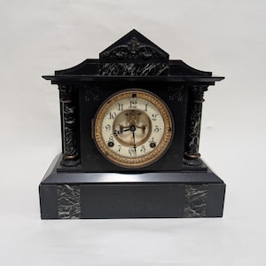 May include: Black antique mantel clock with a gold-toned clock face. The clock has Roman numerals and decorative columns on each side. The clock is made of black marble with white veining.
