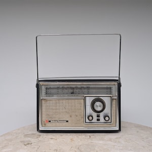 May include: A vintage National Panasonic transistor radio featuring a black rectangular frame, a silver metal carrying handle, a beige speaker grille, and a silver control panel with a large central tuning knob and two smaller volume and tone dials.