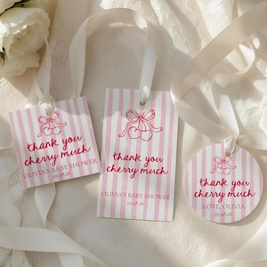 Puede incluir: Tres etiquetas de regalo para baby shower con un diseño a rayas rosas y blancas. Cada etiqueta tiene una ilustración de cerezas y el texto "thank you cherry much". Las etiquetas están atadas con cintas blancas y contienen el texto "OLIVIA'S BABY SHOWER 10.28.26".