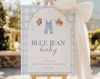 May include: A baby shower sign on an easel with a light blue gingham pattern. The sign reads "BLUE JEAN baby" with illustrations of baby clothes. A large white bow adorns the top right corner. The sign welcomes guests to Olivia's baby shower on 08.24.26.