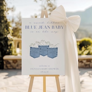 May include: A baby shower welcome sign on a wooden easel. The sign reads "a sweet little BLUE JEAN BABY is on the way." It features three denim pockets with floral accents and the text "welcome to OLIVIA'S BABY SHOWER 08.24.26". A large white bow adorns the easel.