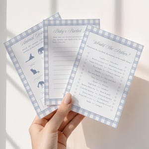 May include: Three baby shower game cards with a blue gingham border. The cards include games like "Animal Guess" and "Would She Rather?" with fill-in-the-blank and multiple-choice questions. The cards are held by a person.