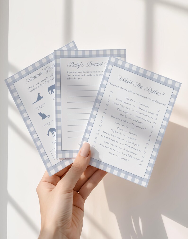 May include: Three baby shower game cards with a blue gingham border. The cards include games like "Animal Guess" and "Would She Rather?" with fill-in-the-blank and multiple-choice questions. The cards are held by a person.