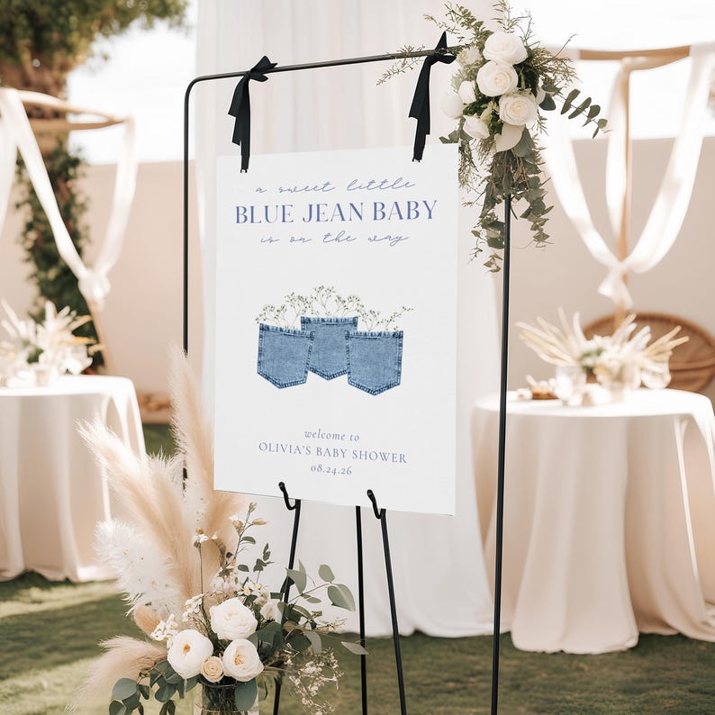 May include: A white sign with the text "a sweet little BLUE JEAN BABY is on the way" and "welcome to OLIVIA'S BABY SHOWER 08.24.26". The sign features three blue jean pockets with floral arrangements. The sign is displayed on a black metal stand.