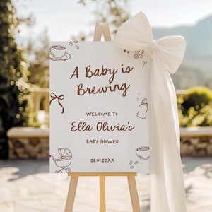 May include: A white sign on a wooden easel with the text "A Baby is Brewing" in brown script. The sign welcomes guests to Ella Olivia's baby shower. A large white bow adorns the top right corner.