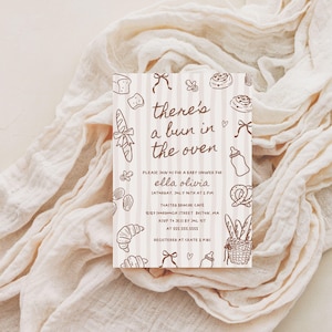 May include: A baby shower invitation with a beige and white striped background. The invitation features brown illustrations of bread, bows, and baby items. Text reads "there's a bun in the oven" and details for the event.