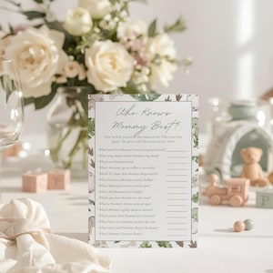 Puede incluir: Una tarjeta de juego para baby shower con el texto "Who Knows Mommy Best?" se muestra en una mesa. La tarjeta tiene un tema de dinosaurios con un arreglo floral en el fondo. Otras decoraciones incluyen un tren de juguete y peque&ntilde;as esferas rosas y grises.