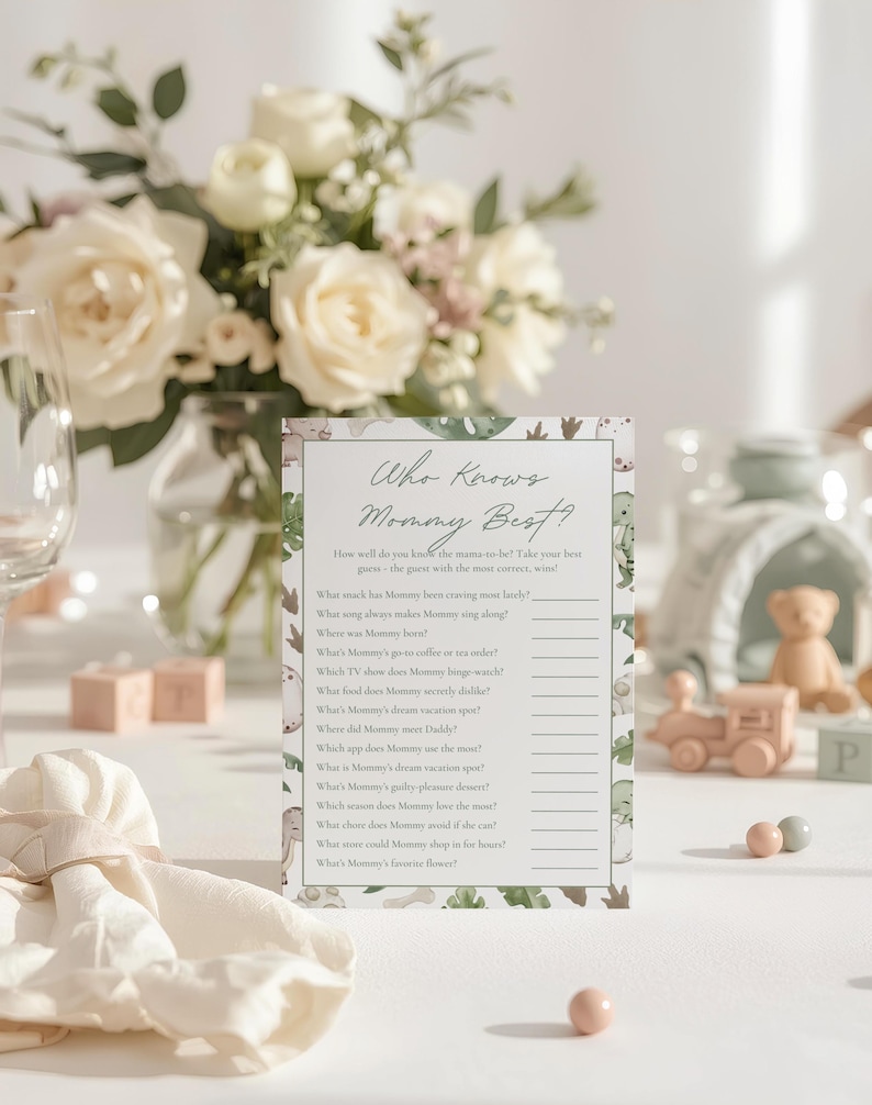 Puede incluir: Una tarjeta de juego para baby shower con el texto "Who Knows Mommy Best?" se muestra en una mesa. La tarjeta tiene un tema de dinosaurios con un arreglo floral en el fondo. Otras decoraciones incluyen un tren de juguete y peque&ntilde;as esferas rosas y grises.