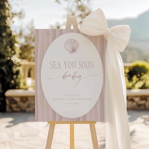 Puede incluir: Un caballete de madera exhibe un letrero de baby shower con el texto "SEA YOU SOON baby". El letrero tiene un gráfico de concha y está decorado con un gran lazo blanco. El letrero da la bienvenida a los invitados al baby shower de Olivia.