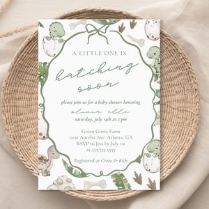 Può includere: Un invito a un baby shower a tema dinosauri. L'invito presenta un bordo verde con illustrazioni di dinosauri e il testo "A Little One Is Hatching Soon". L'invito include i dettagli per un baby shower in onore di Olivia Elle.