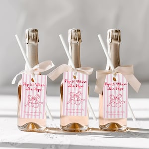 May include: Three champagne bottles with pink liquid, each adorned with a pink and white striped tag that reads "Pop it When She Pops." The tags feature a cherry design and are tied with a light pink ribbon. White straws are inserted.