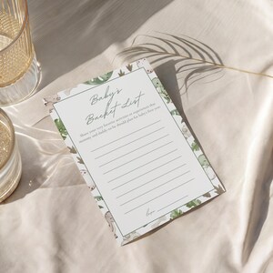 Puede incluir: Una tarjeta blanca de "Baby's Bucket List" con un borde verde de dinosaurios y hojas. La tarjeta tiene l&iacute;neas para escribir y el texto "Baby's Bucket List" en escritura cursiva. La tarjeta est&aacute; sobre una superficie de tela blanca.