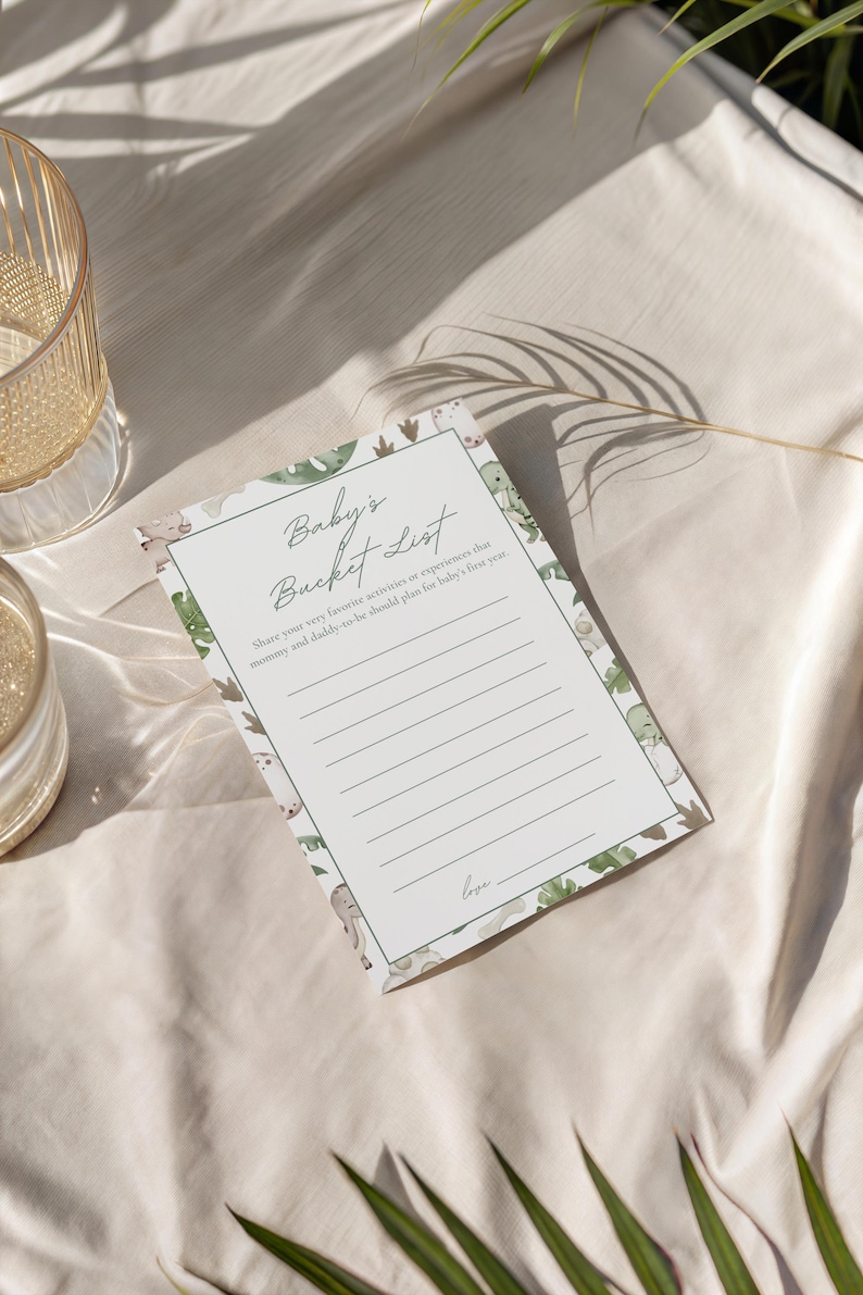 Puede incluir: Una tarjeta blanca de "Baby's Bucket List" con un borde verde de dinosaurios y hojas. La tarjeta tiene l&iacute;neas para escribir y el texto "Baby's Bucket List" en escritura cursiva. La tarjeta est&aacute; sobre una superficie de tela blanca.